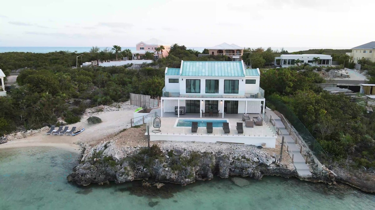 The two-level villa is viewed from a distance, highlighting its modern design and expansive outdoor pool area. Surrounding greenery complements the ocean, with a private beach cove visible nearby. The structured layout includes large glass doors, inviting light and a seamless connection to the landscape.