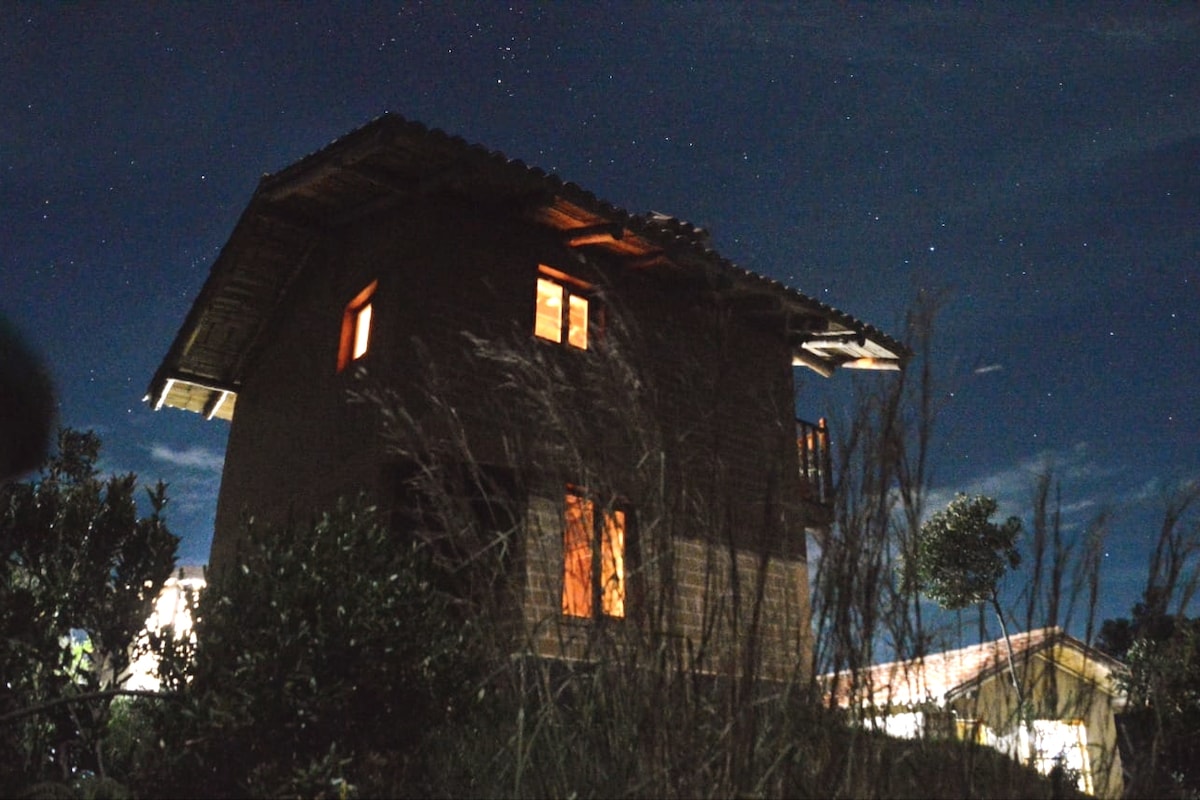 A charming two-story cabin is shown illuminated against a night sky filled with stars. Soft light emanates from the windows, providing a glimpse of its inviting interior, while tall grass surrounds the base, enhancing the sense of tranquility and connection to nature.