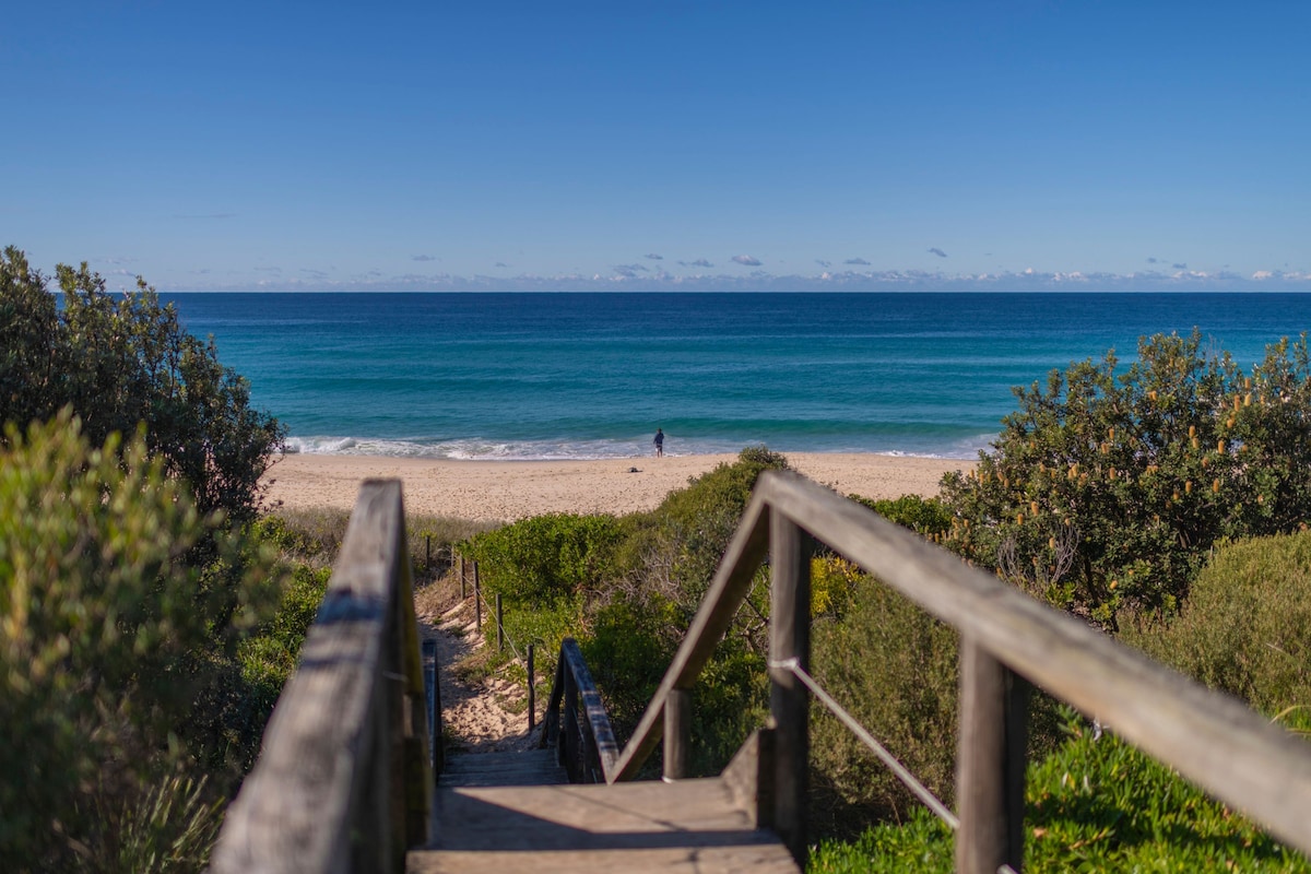 A pathway leads from a wooden staircase down to the sandy beach, framed by lush greenery on either side. The gentle waves of the ocean glisten under clear blue skies, inviting relaxation and exploration.