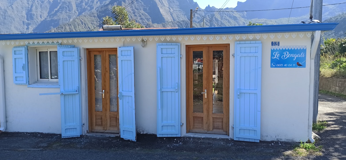 A charming building with light blue shutters and wooden doors is pictured. The structure features a sign labeled 'Le Bungalow.' Lush mountainous scenery provides a stunning backdrop, highlighting the peaceful surroundings. An organized entrance leads to multiple access points of the accommodation.