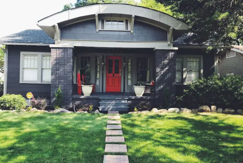 The charming exterior of a grey house is framed by lush green grass and well-maintained landscaping. A stone walkway leads to a double door entrance painted in a vibrant red, flanked by matching red chairs and decorative potted plants.