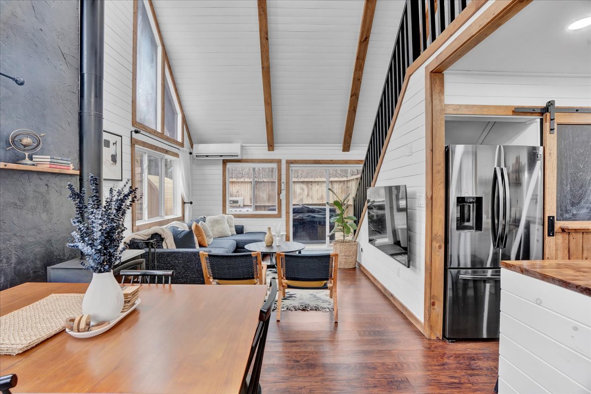 An open living space features high, vaulted ceilings with wooden beams. A modern seating area is positioned next to large windows, allowing natural light to fill the room. A dining table is visible, alongside an efficient kitchen equipped with stainless steel appliances.