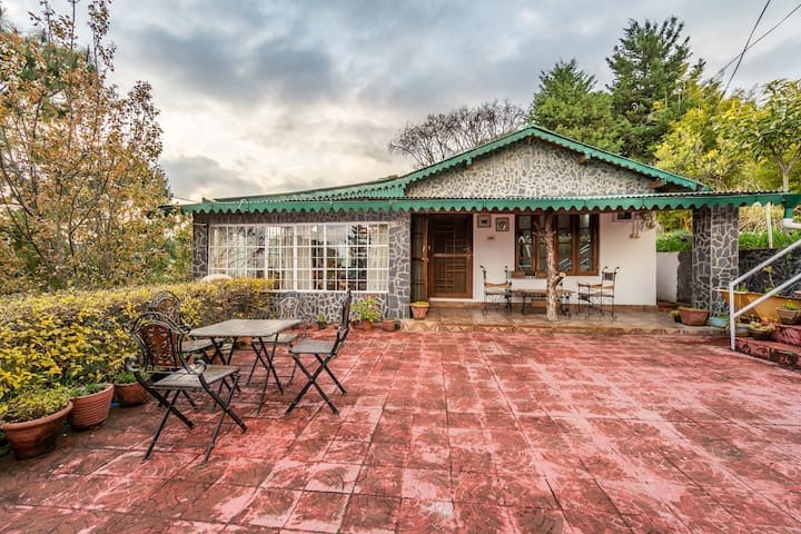The exterior of the cozy cabin features a stone facade with a green roof, surrounded by lush greenery. A spacious patio, adorned with potted plants, showcases a traditional wooden bench and two tables with chairs, creating an inviting area for relaxation.