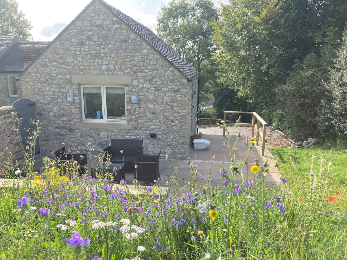 A charming limestone holiday home is positioned on a hillside, surrounded by vibrant wildflowers. The property features a private patio that opens up to the countryside. The natural landscape includes lush greenery and a variety of flowering plants, enhancing the outdoor setting.