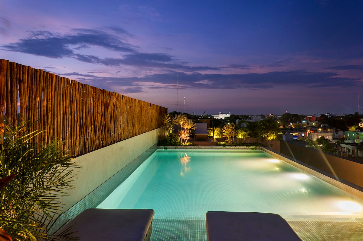 A serene rooftop area is highlighted by a large coral pink pool, surrounded by palm trees and wooden fencing. The evening sky provides a soft backdrop, enhancing the tranquil atmosphere and offering views of the Tulum skyline.