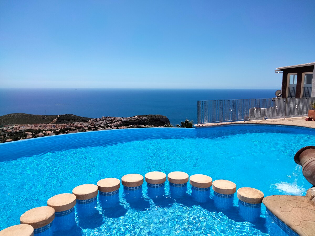 An infinity pool with inviting turquoise water overlooks the Mediterranean Sea. Stone steps lead into the pool, offering a seamless transition to the stunning views of the coastline and horizon under a clear blue sky.