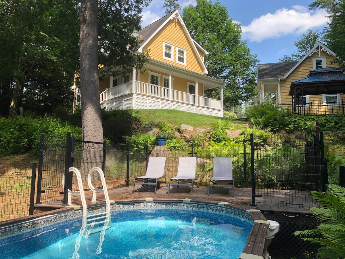 A tranquil outdoor space features a heated swimming pool surrounded by a security fence. Lounge chairs are positioned beside the pool, with a backdrop of lush greenery and a charming yellow house visible in the distance.