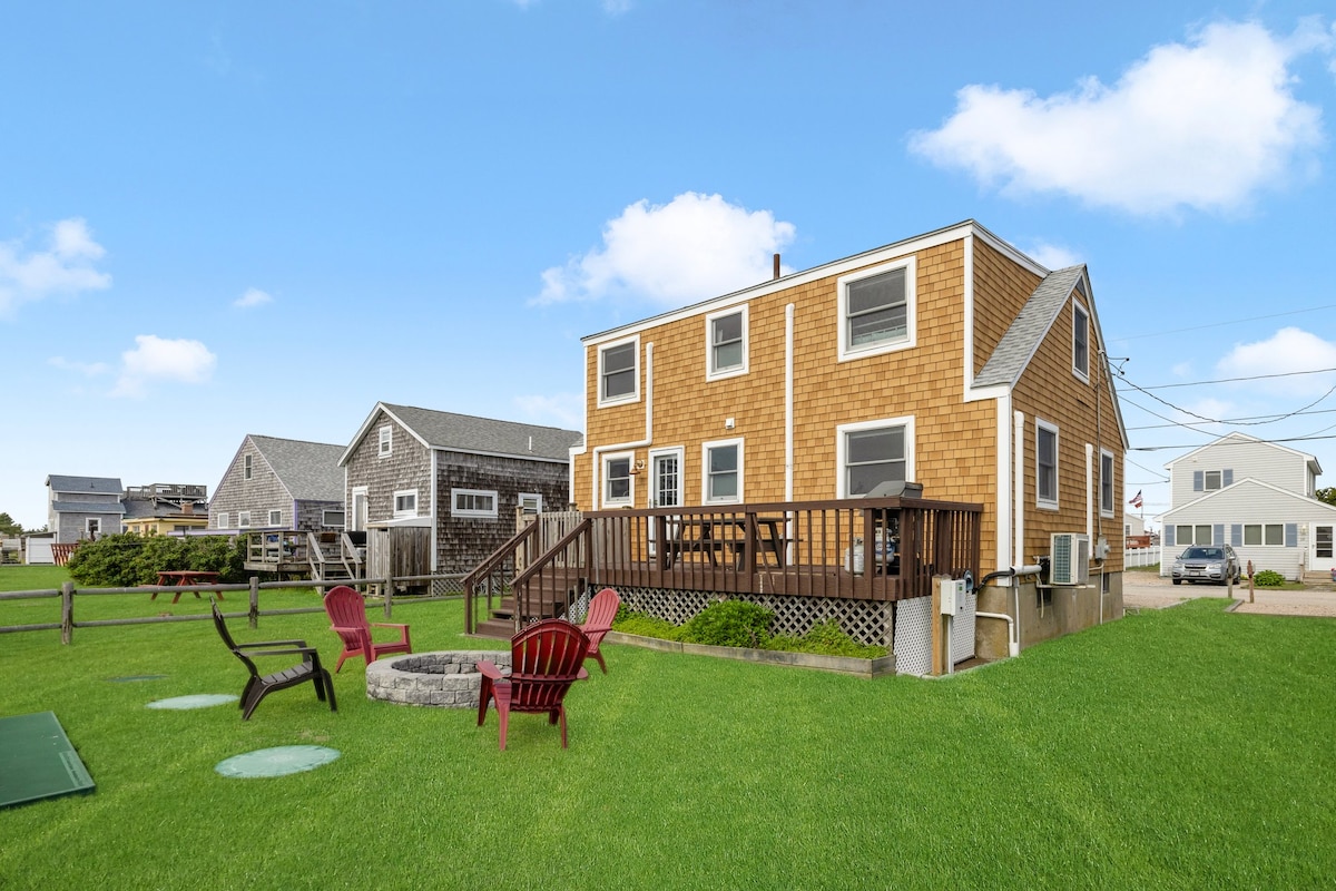 A charming two-story cottage is showcased, featuring a wooden deck with entry steps. Surrounding the structure, an outdoor fire pit with red chairs is set on a green lawn, while neighboring cottages can be seen in the background against a clear blue sky.