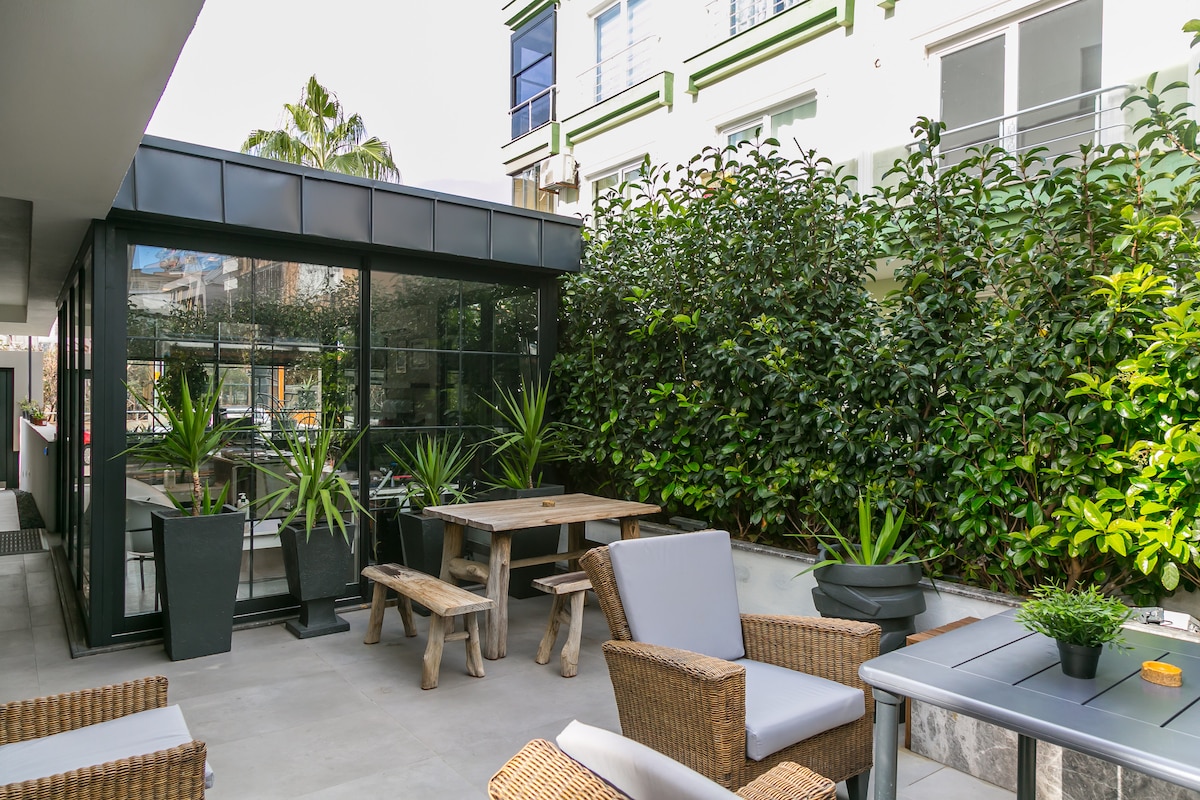 An outdoor seating area features wooden tables and comfortable chairs surrounded by lush greenery. Glass walls of the adjacent building reflect the foliage, creating a serene environment for relaxation. Potted plants add to the inviting atmosphere, contributing to an overall sense of tranquility.