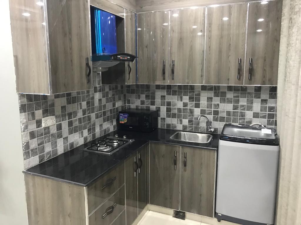 A compact kitchen features sleek cabinetry in shades of gray. Glass tiles create a modern backsplash, while appliances include a refrigerator, washing machine, and microwave. A gas stove is positioned next to a stainless steel sink, providing functional kitchen amenities in a clean, organized layout.