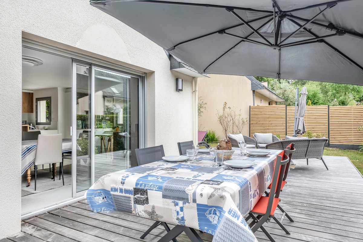 An outdoor terrace is depicted with a large dining table set for a meal, complemented by a durable umbrella providing shade. Surrounding chairs are arranged for seating. The scene showcases a seamless connection to the interior space, visible through sliding glass doors.