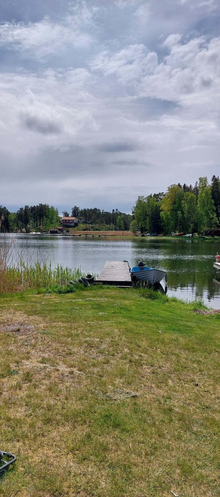 A serene lakeside view captures a wooden dock extending into calm waters. Lush greenery surrounds the lake, with trees lining the shore. In the distance, a house is visible, harmonizing with the natural setting.