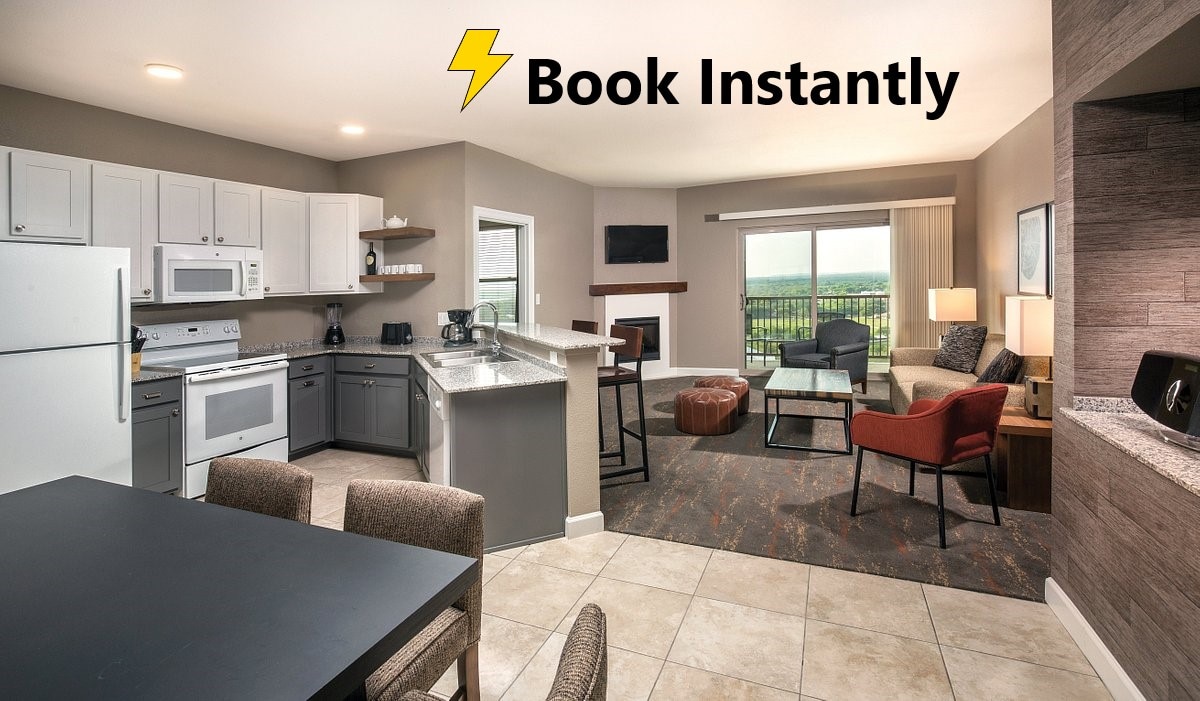 A modern kitchen is paired with a dining area featuring a dark table and light-colored chairs. The living space includes a cozy seating arrangement with a sofa, armchairs, and a coffee table. Natural light fills the room through large windows, showcasing views of the surrounding hills.