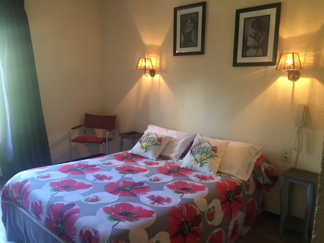 A sleeping area features a spacious bed adorned with floral-patterned bedding and contrasting pillows. A stylish chair stands to the side, while soft lighting from wall sconces creates a warm atmosphere. Artwork hangs above the bed, enhancing the room's character.