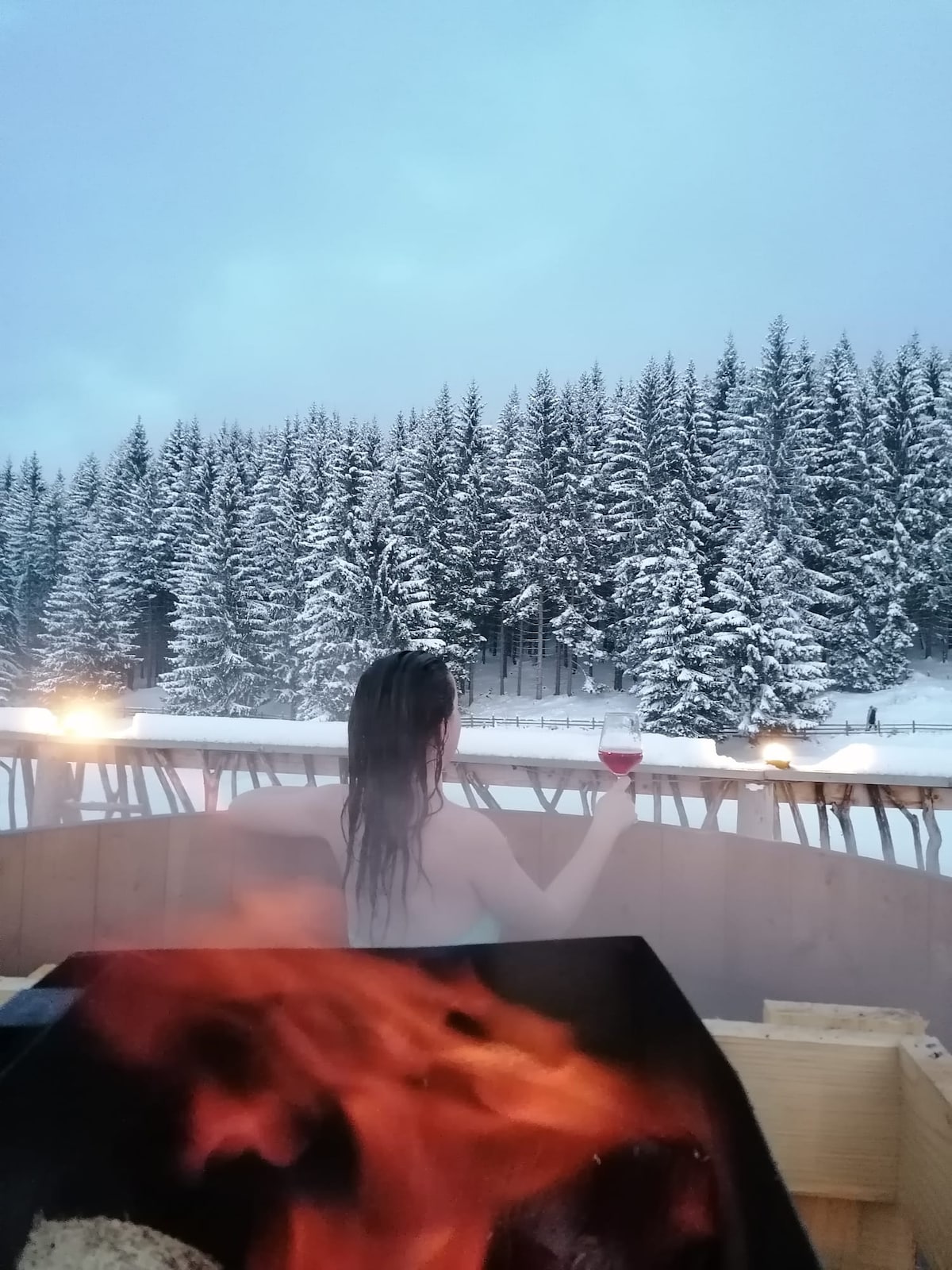 A wooden hot tub is shown in a snowy landscape, with steam rising in the air. A person, viewed from behind, enjoys a drink while overlooking a dense forest of snow-covered trees in the distance.