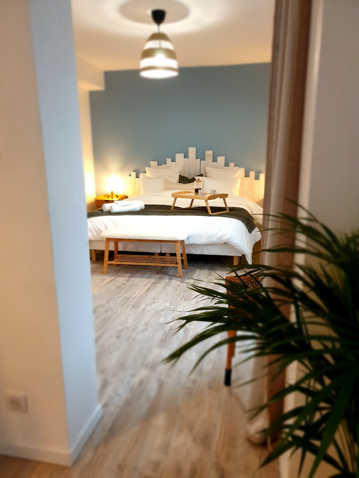 A spacious bedroom features a king-size bed adorned with soft linens and a decorative headboard. A small table with a tray is positioned at the foot of the bed. Natural light fills the room, complemented by a soothing blue wall and wooden flooring.