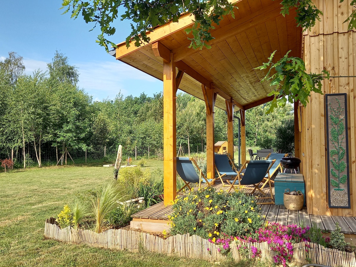A wooden terrace is covered and faces a garden with colorful flowers and greenery. Two lounge chairs are positioned under the structure, providing a relaxed seating area. In the background, trees line the edge of the yard, enhancing the serene environment.
