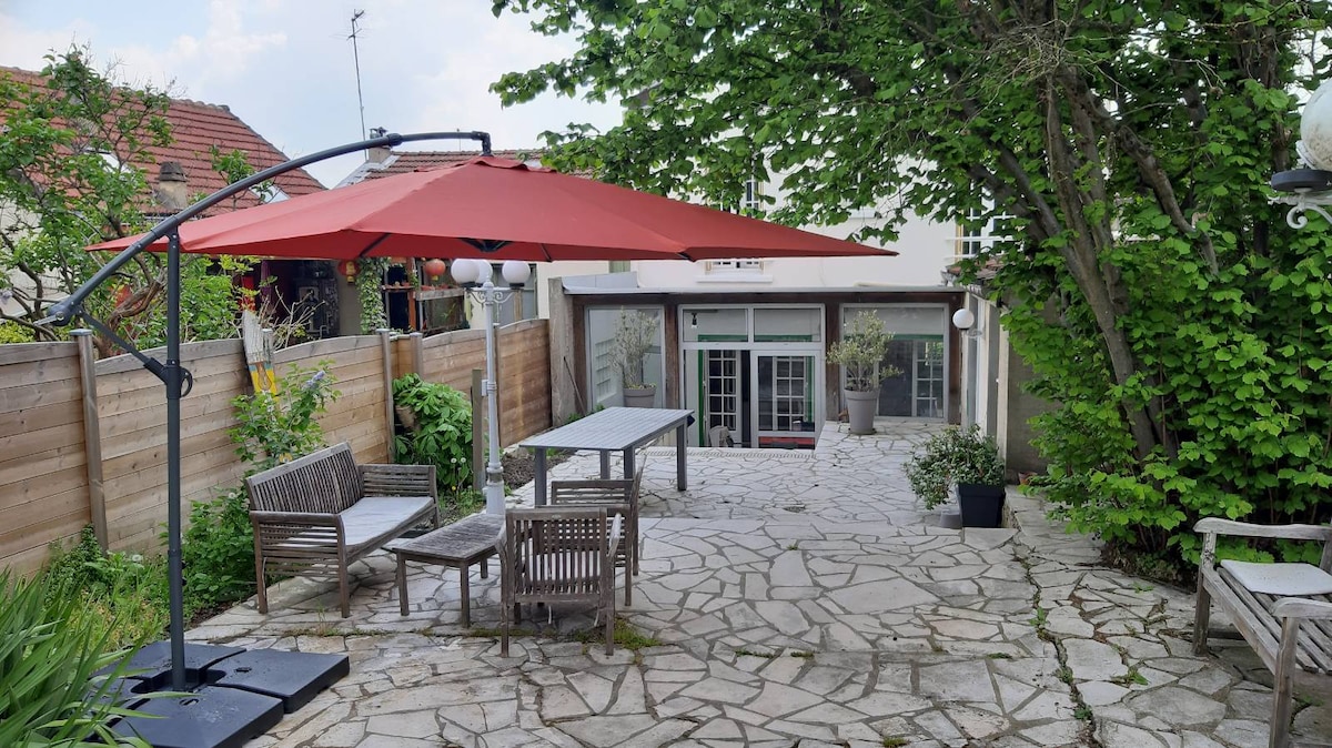 An outdoor patio space is presented, featuring a sturdy table and several wooden chairs under a large red umbrella. Natural stone flooring is visible, surrounded by greenery and garden features. A cozy area for relaxation is created in this inviting and well-maintained garden.