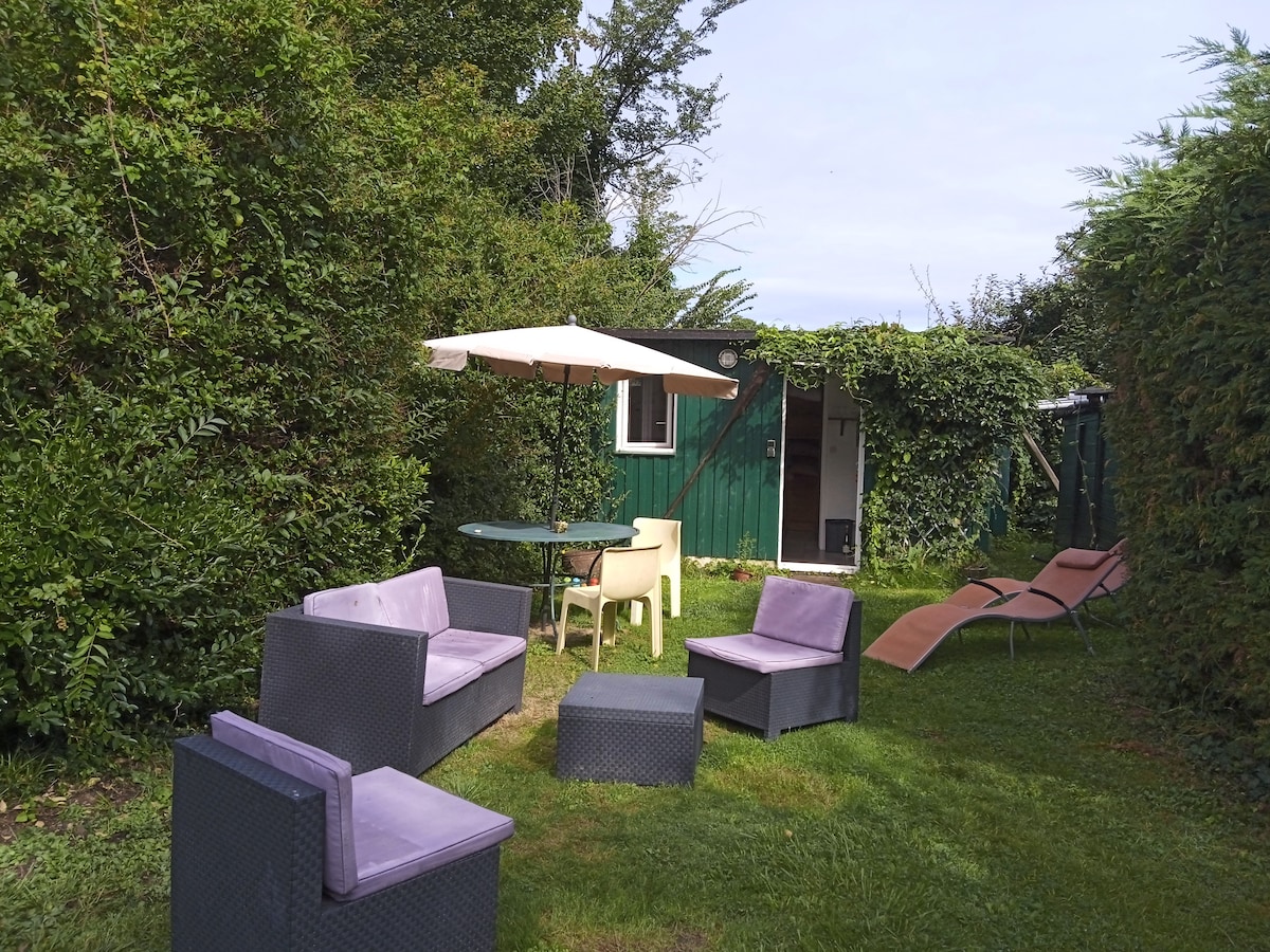 A private outdoor garden area is surrounded by greenery, featuring a table with four chairs and two lounge chairs. A parasol provides shade over the seating arrangement, while a small green structure can be seen in the background, indicating the entrance to the studio.