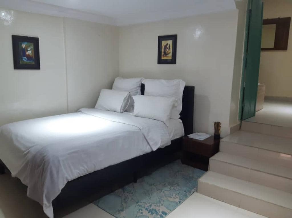 A spacious bedroom features a neatly made bed with white linens and decorative pillows. Two framed artworks are displayed on the walls. Natural light illuminates the room, highlighting the light-colored decor and the tiled flooring leading to steps that ascend to another area.