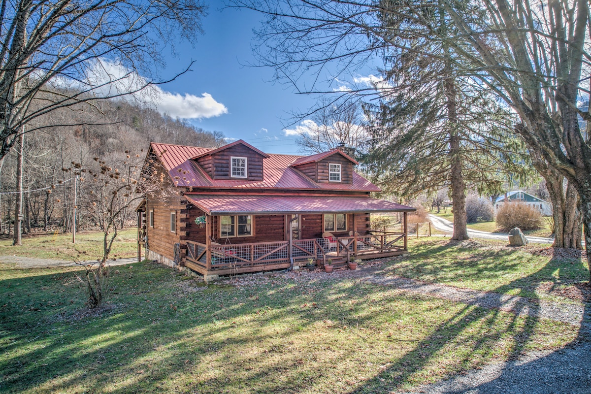 A log cabin with a red metal roof is set on a grassy area surrounded by trees. A spacious front porch features seating, and the structure displays multiple windows that allow natural light to enter, accentuating the cozy mountain retreat ambiance.