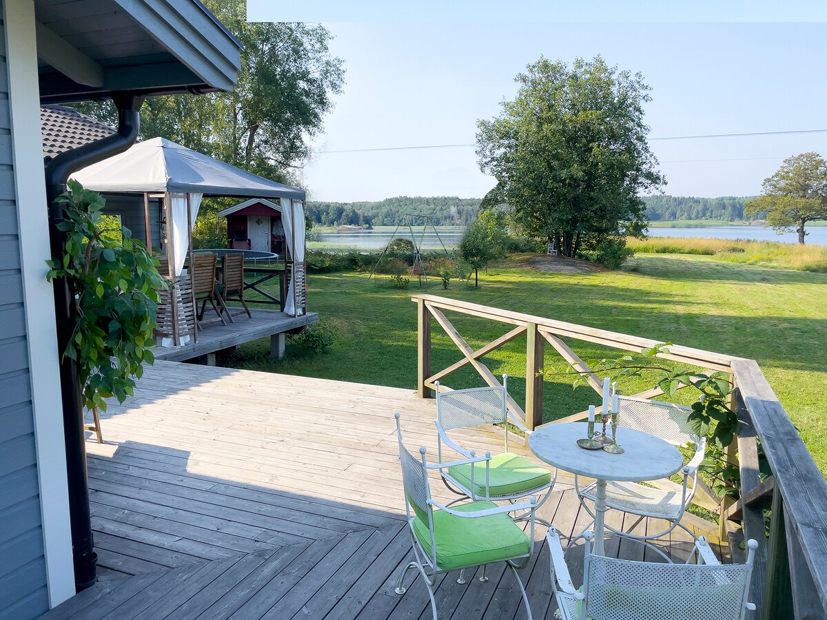 A spacious wooden deck features a round table and chairs, complemented by green cushions. A gazebo is visible in the background, with a view of a scenic lake and expansive green fields. The area offers a tranquil setting for outdoor relaxation.