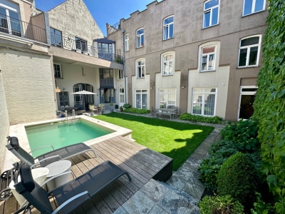 A serene courtyard is presented, showcasing a small pool surrounded by wooden decking and well-manicured grass. Comfortable lounge chairs are positioned around the pool, while garden seating is arranged nearby. The surrounding building features large windows that allow natural light to illuminate the area.