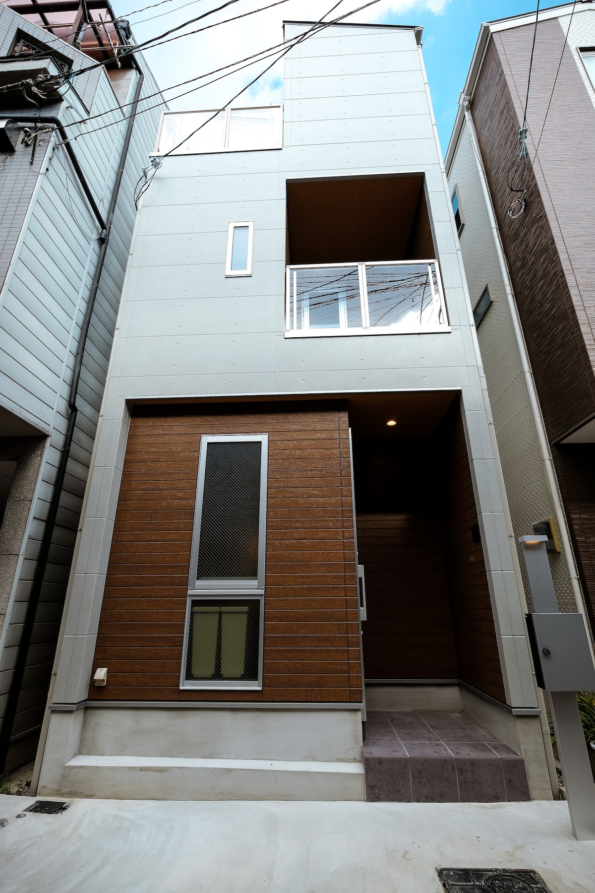 The exterior of a modern, multi-story building features a combination of sleek metal and warm wooden paneling. A large entrance area leads up to expansive windows on the upper floor, while the ground level includes a small porch with a stone pathway.