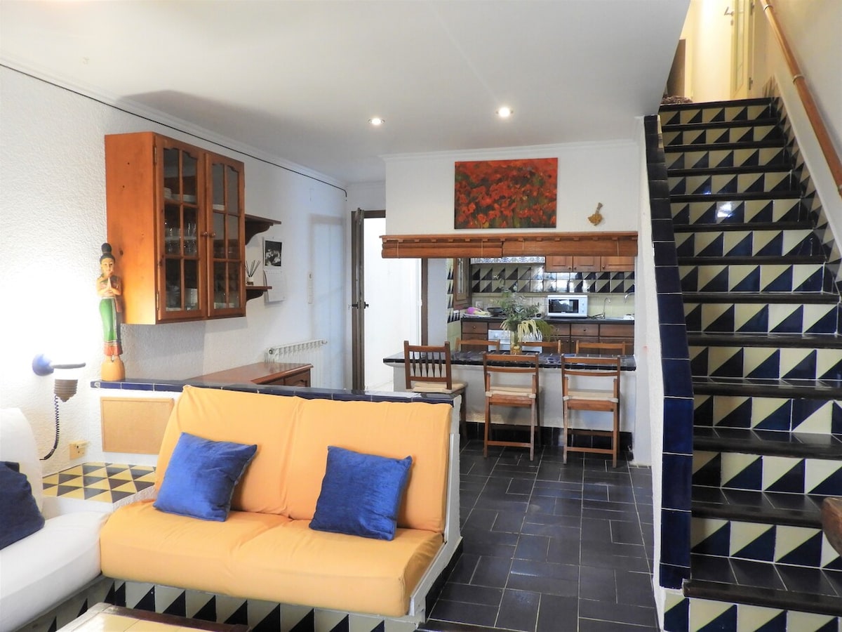 The interior showcases a cozy lounge area featuring a yellow sofa with blue cushions. A wooden cabinet is mounted on the white wall, and the kitchen area has a dining table set for four. The stairs lead to the upper level, highlighted by a patterned tile design.