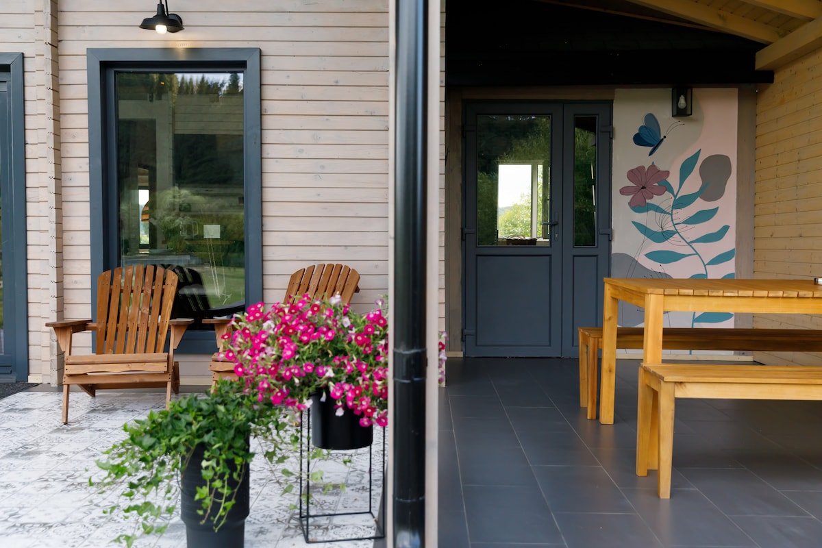 An outdoor seating area features two wooden chairs placed on a tiled patio, surrounded by blooming potted plants. The adjoining space showcases a simple wooden table and colorful wall art, enhancing the serene environment of the chalet.