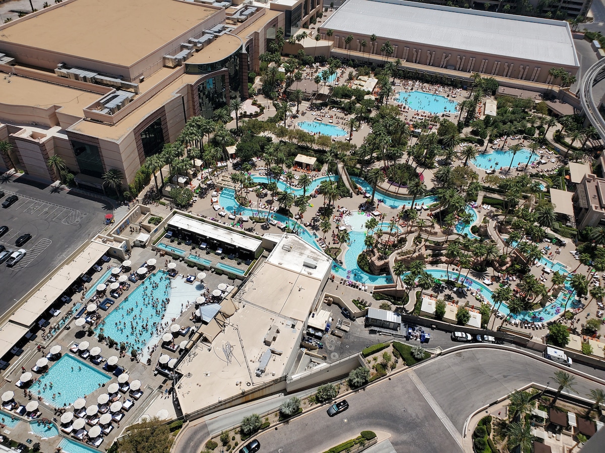 An aerial view captures a sprawling pool area, featuring a lazy river winding through lush palm trees and sun loungers. Numerous guests enjoy the sparkling water, with various amenities visible, including shaded cabanas and a bar area. The surrounding resort structure adds to the vibrant atmosphere.