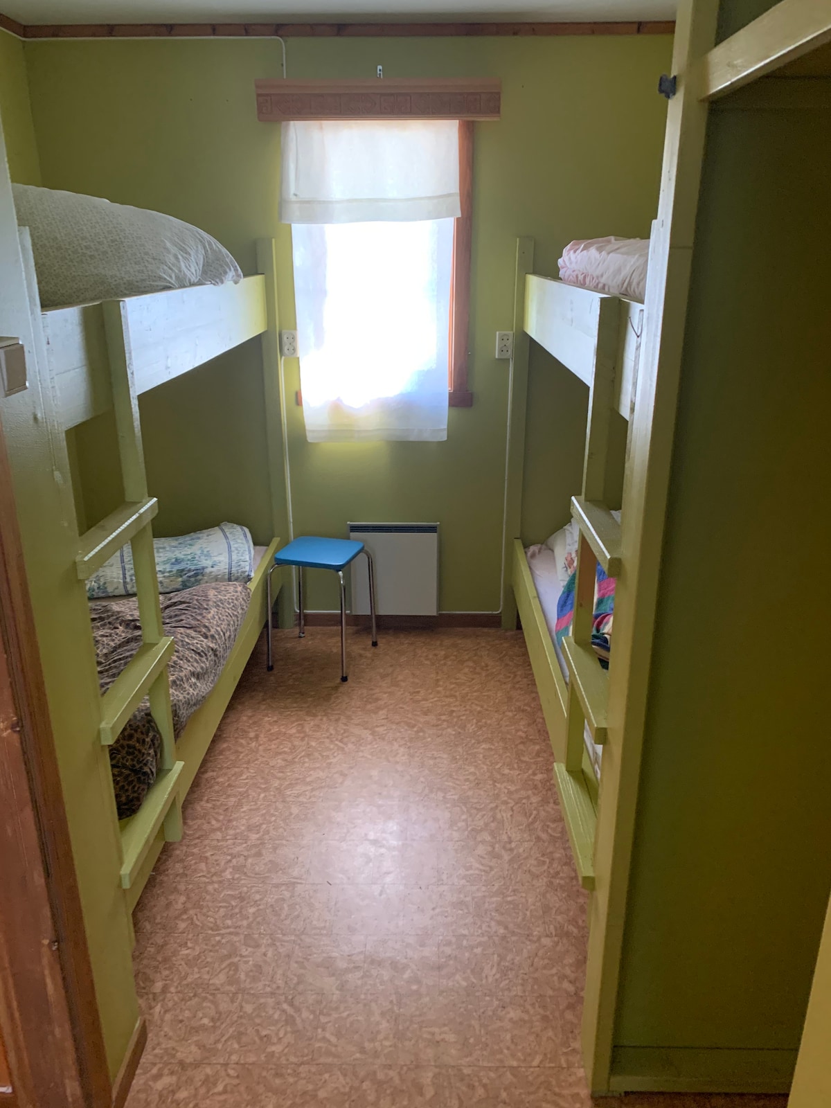 A small room with green walls features two sets of bunk beds, each with a neatly arranged bedding. A simple blue stool is positioned between the bunks. Natural light enters through a window partially covered by a light curtain, illuminating the space.