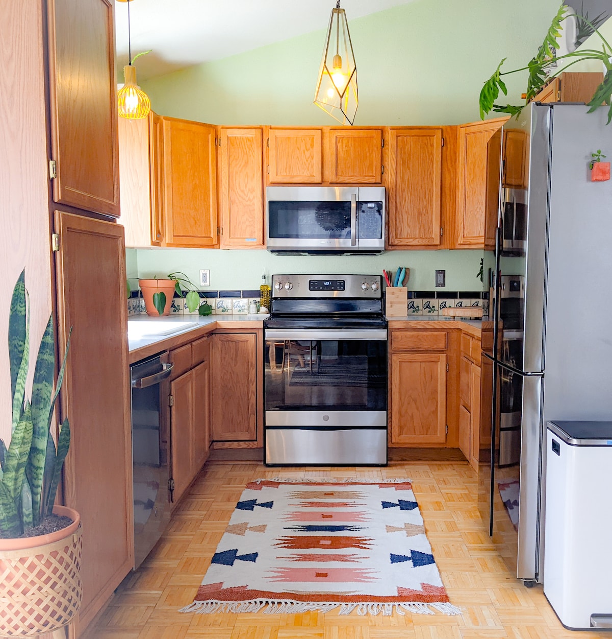 A well-equipped kitchen features wooden cabinets and stainless steel appliances. A patterned area rug adds a touch of warmth to the hardwood floor. Green plants are seen on the countertop, enhancing the bright and inviting atmosphere of the space.