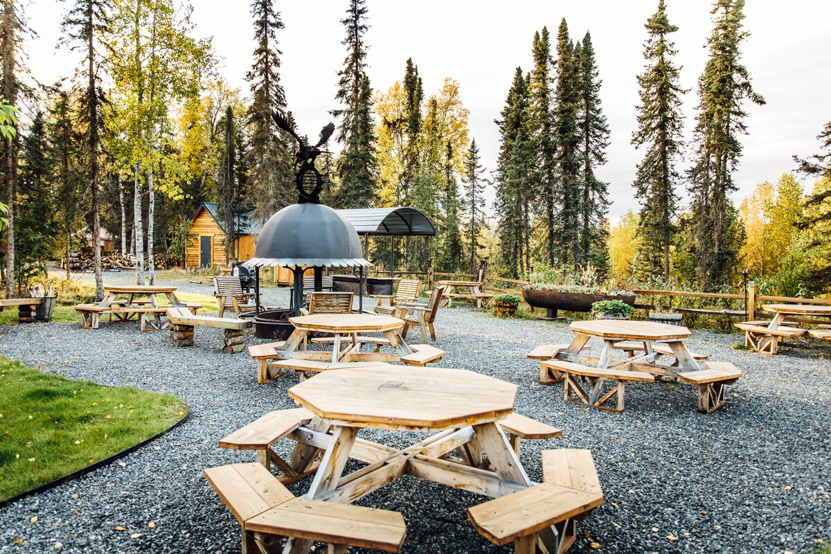 An outdoor gathering area is surrounded by tall, leafy trees. Multiple hexagonal wooden tables with benches are arranged across a gravel surface. A large, domed fire pit is positioned centrally, providing a communal space for guests to enjoy.