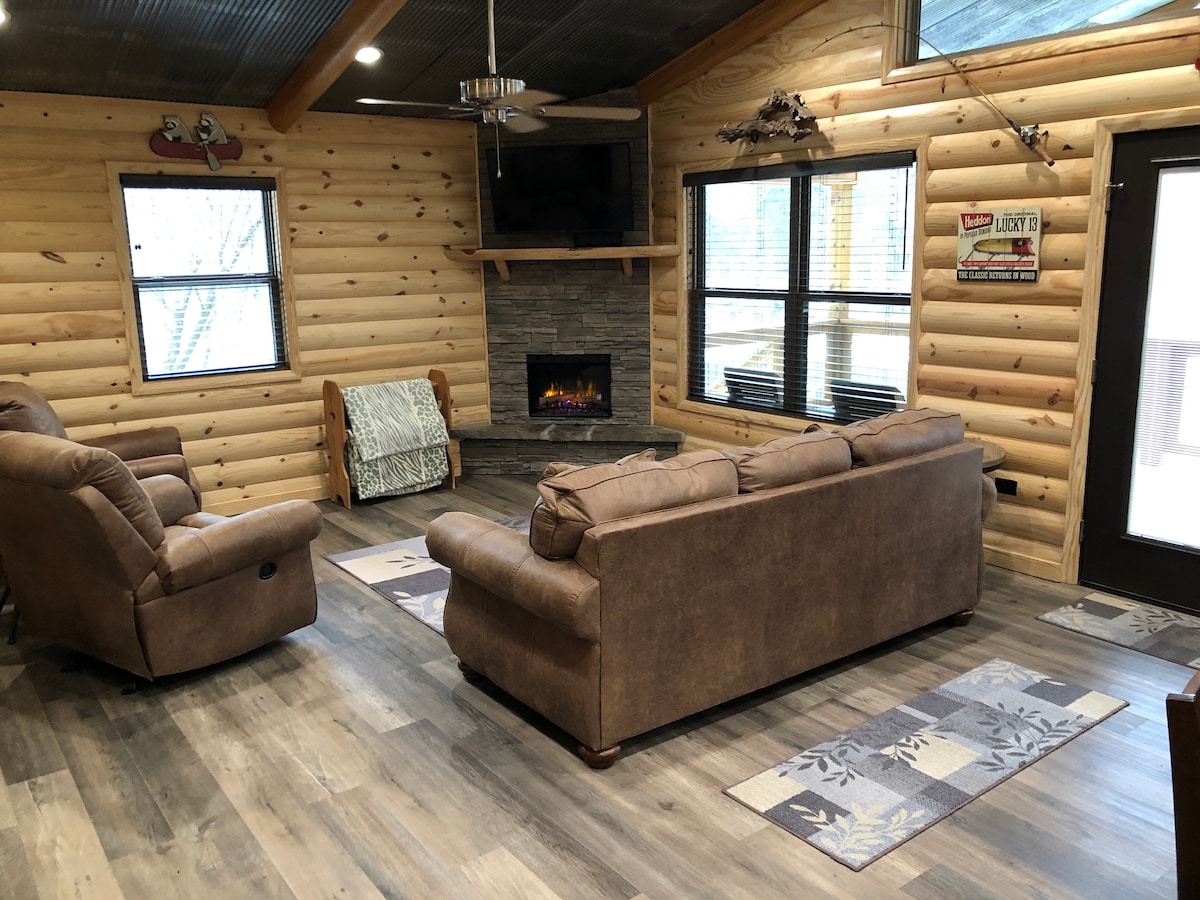 A welcoming living space features a cozy, neutral-toned sofa and reclining chairs. A stone fireplace is situated below a television, providing a focal point for the area. Large windows allow ample natural light, illuminating the wood-paneled walls and comfortable furnishings.