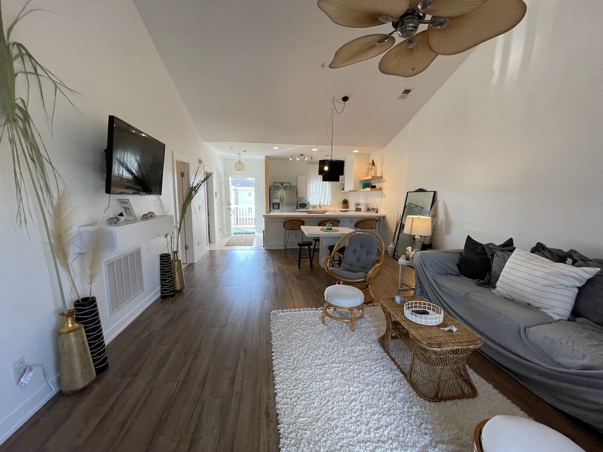 A modern living area is highlighted by a spacious gray sofa and decorative chairs. A wall-mounted TV is positioned above a minimalist fireplace. Textured rugs cover the floor, while natural light floods the space through large windows, enhancing the open atmosphere.