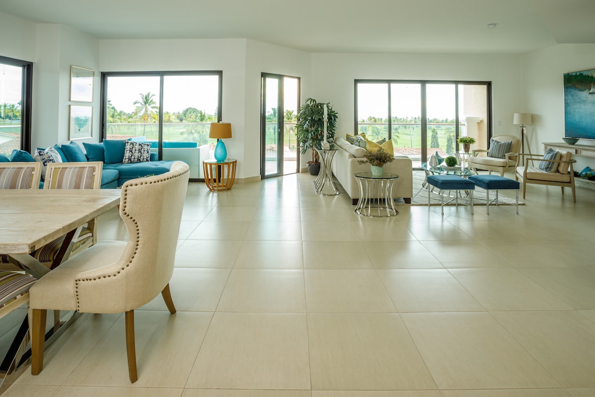 The living area features a spacious layout with natural light flowing through large windows. A comfortable blue sectional couch and neutral armchairs are arranged around a coffee table. A dining table with upholstered chairs occupies one corner, while a scenic view of greenery is visible through the glass doors.