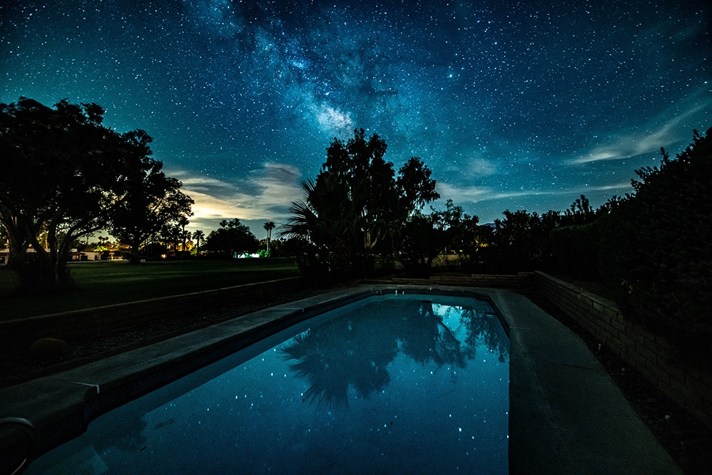 A serene view of a swimming pool under a star-filled night sky, with the Milky Way reflecting on the still water. Surrounding trees and shrubs frame the pool area, providing a sense of seclusion amidst the natural landscape.