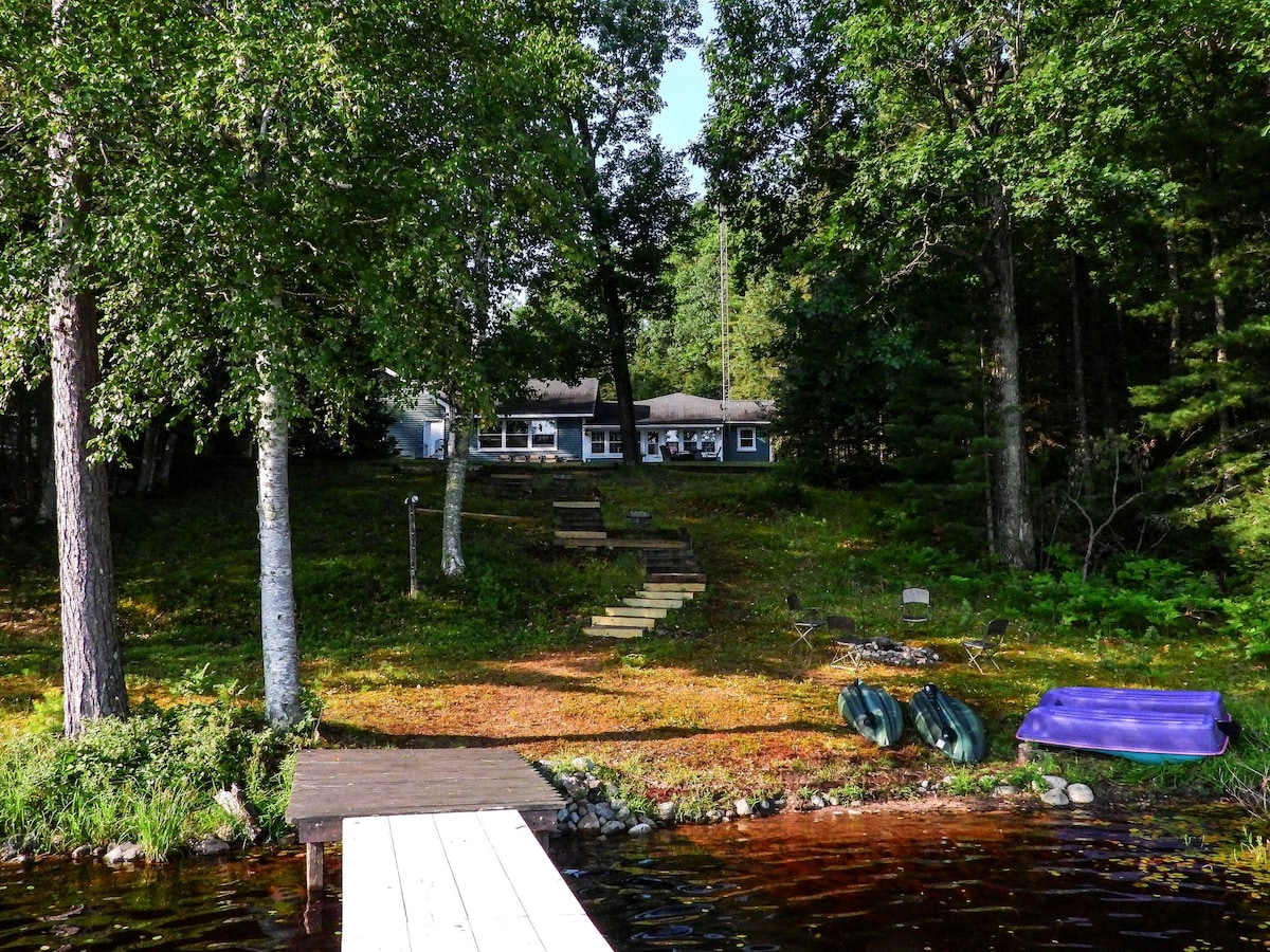 A multi-level access leads from the water's edge to the welcoming home nestled among trees. The vibrant green grass and bushes surround the property, with a wooden dock visible in the foreground. Relaxation equipment, including kayaks and a paddleboat, is situated near the water.