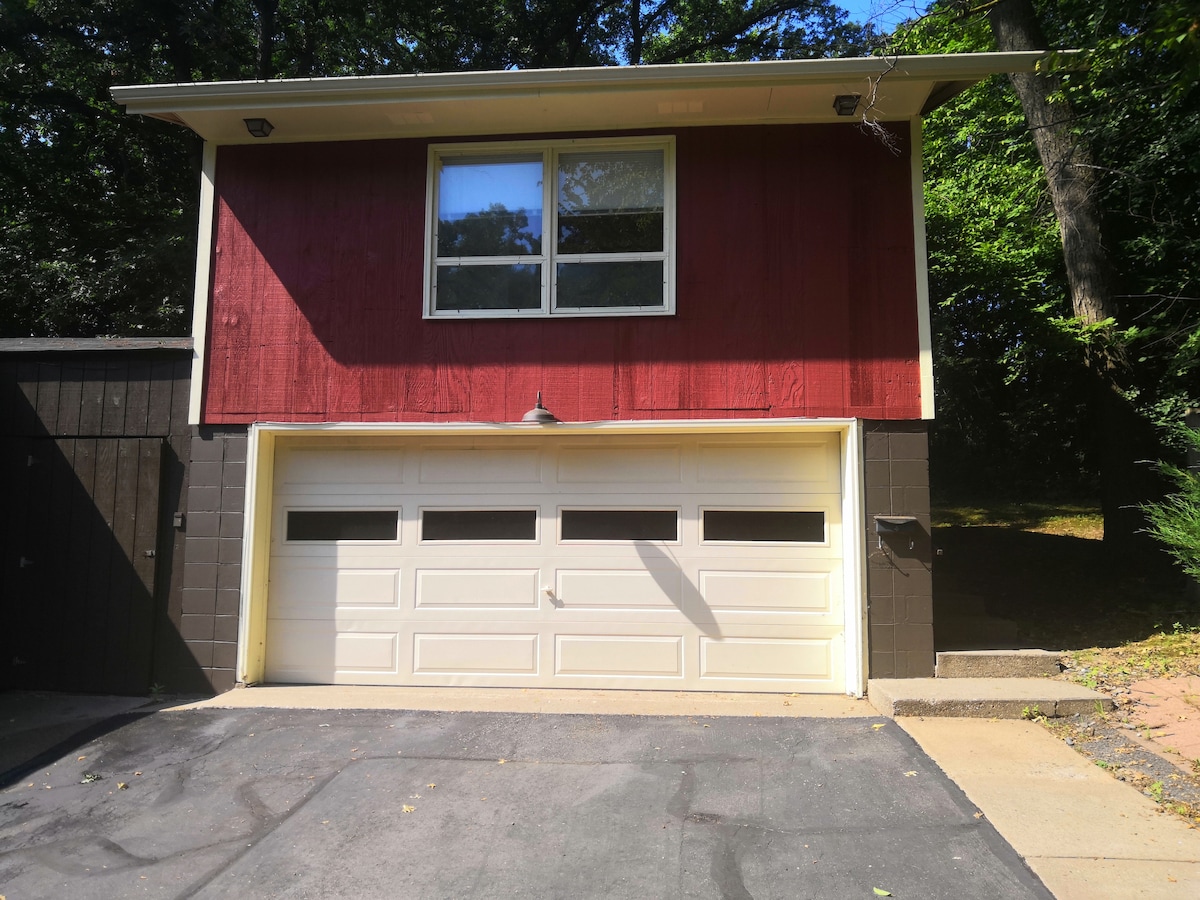 The exterior of the space showcases a two-story structure with a striking red wooden facade and large windows. A three-car garage door in a light color is visible, framed by a neatly paved driveway leading to a wooded area.