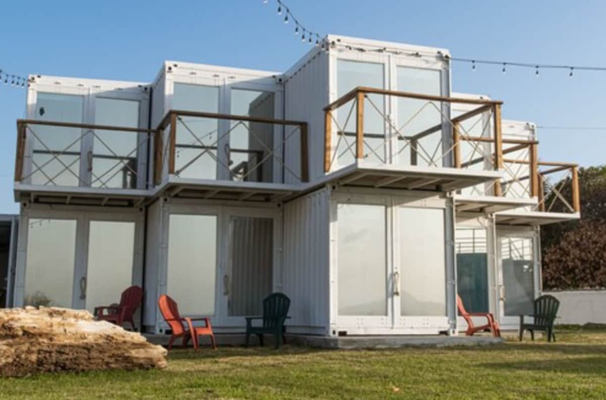 A modern building constructed from shipping containers features large glass windows and multiple balconies. Colorful chairs are positioned in front on a grassy area, providing seating options for guests. String lights are draped overhead, enhancing the outdoor space.