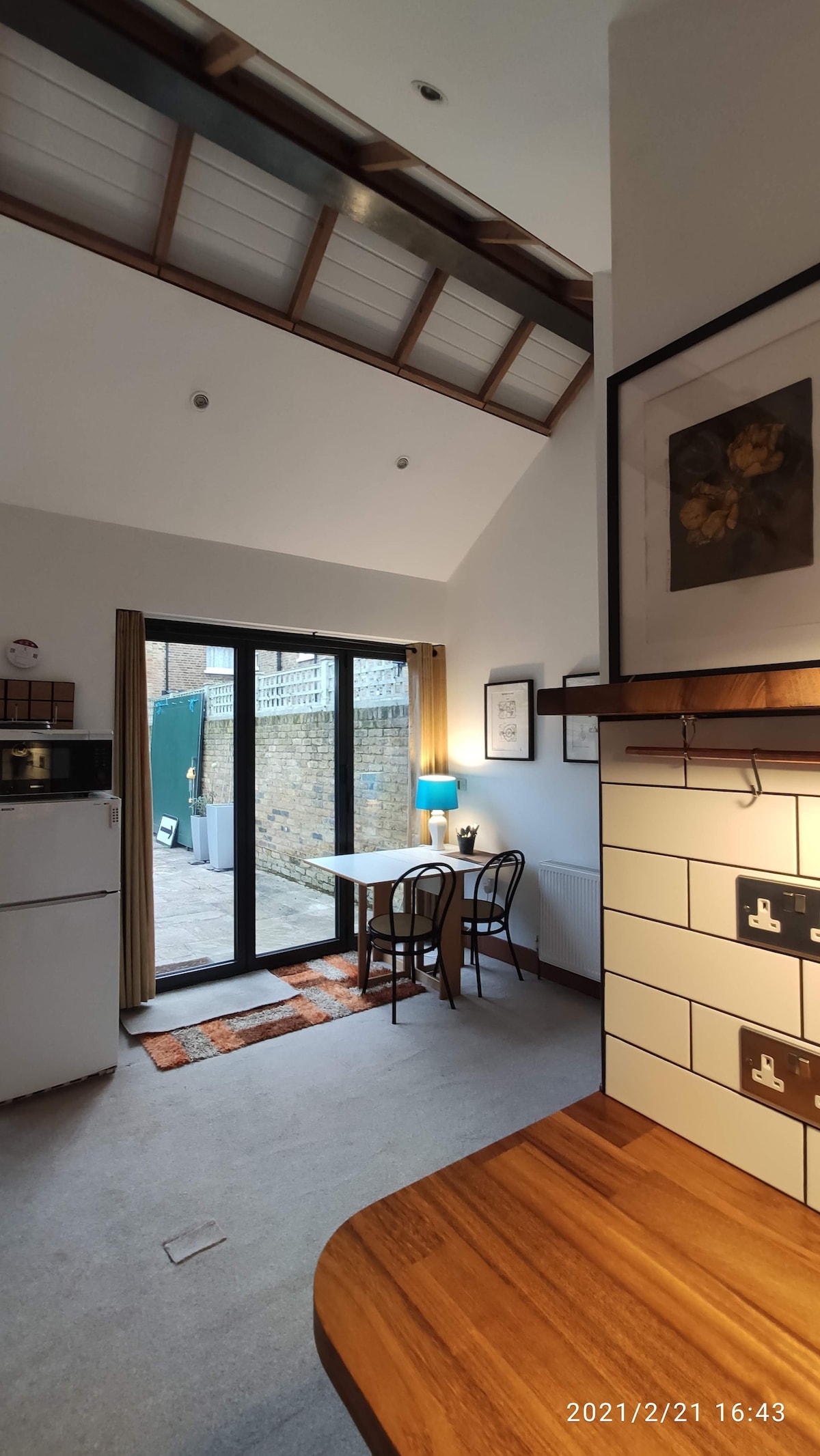The image showcases a kitchen area with wooden countertops and white cabinetry. A dining table with two chairs is positioned next to bi-fold doors that open to the courtyard. Natural light floods the space, highlighting the high arched ceiling and neutral color scheme.