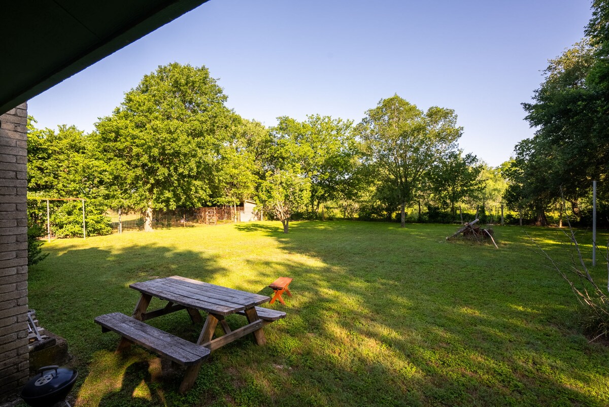 A spacious backyard is surrounded by lush trees, providing a natural setting. A wooden picnic table sits on well-maintained grass, offering a space for outdoor dining. A fire pit is visible in the area, perfect for evening gatherings under the open sky.