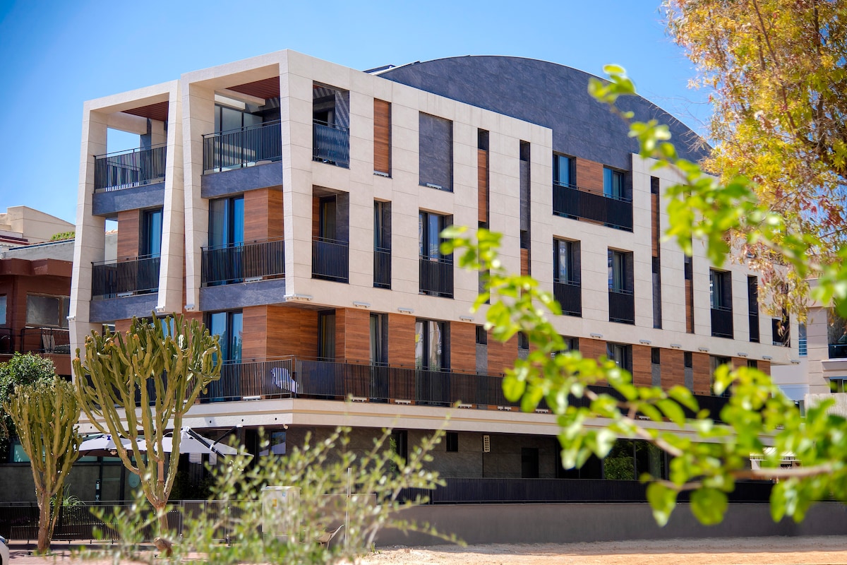 A modern building is shown in a blend of light-colored stone and wood finishes, featuring multiple balconies with black railings. Surrounding greenery includes low shrubs and cacti, contributing to the inviting exterior. The structure is complemented by a clear blue sky.