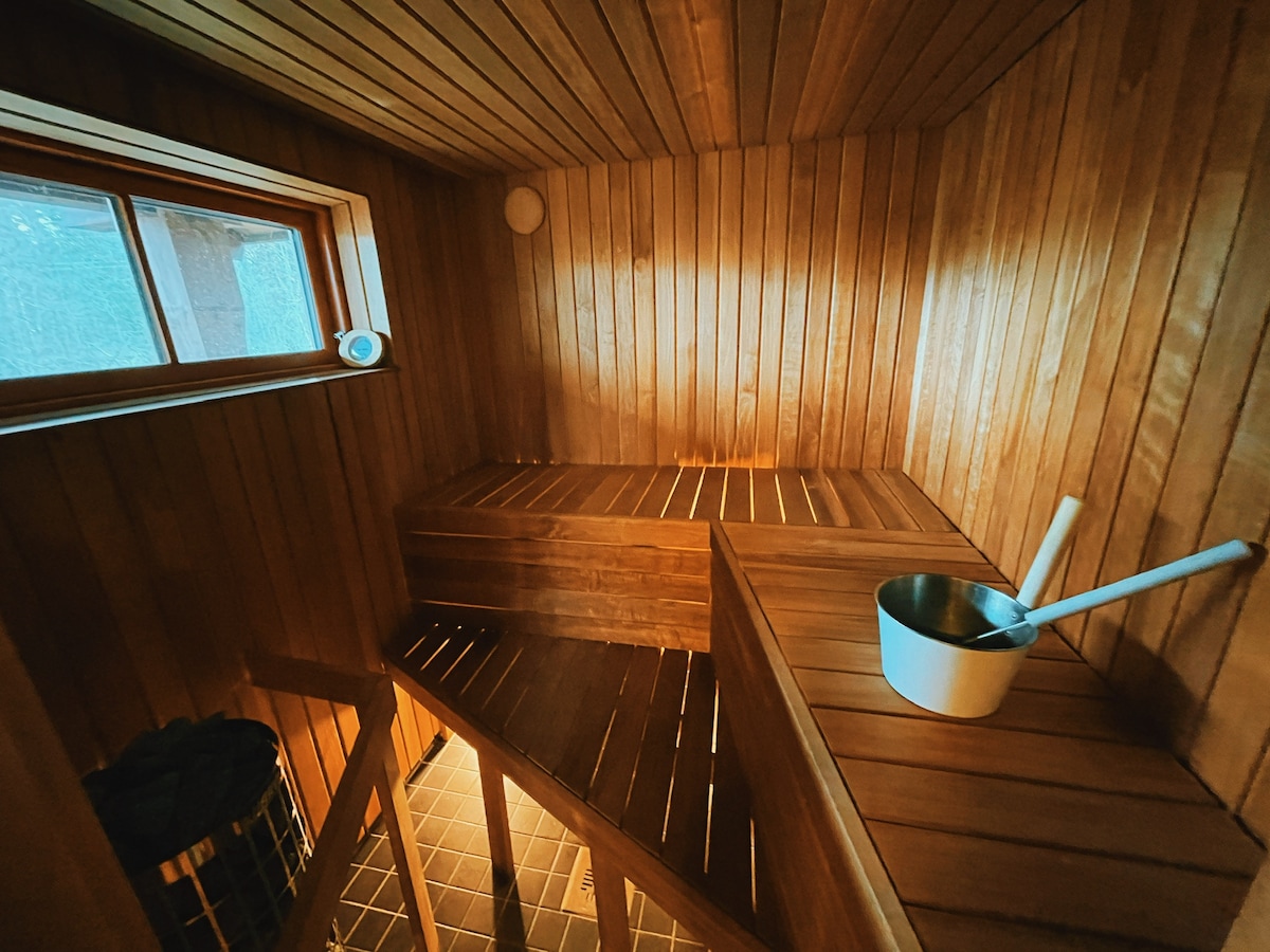 The sauna features wooden walls and benches, with a small window allowing natural light. A metal bucket and ladle are placed on the bench. The tiled floor provides a contrasting texture, while soft lighting creates a calming ambiance.