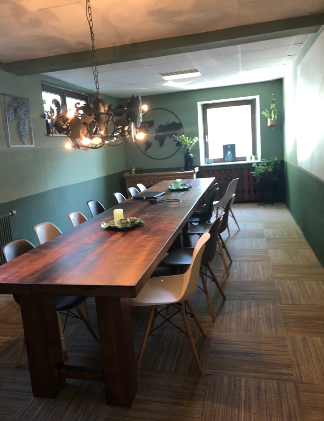A long wooden dining table filled with decorative items is surrounded by a mix of seating options. The room features green walls, plants placed along the windowsill, and light fixtures that add warmth. Natural light filters in through the windows, enhancing the space.