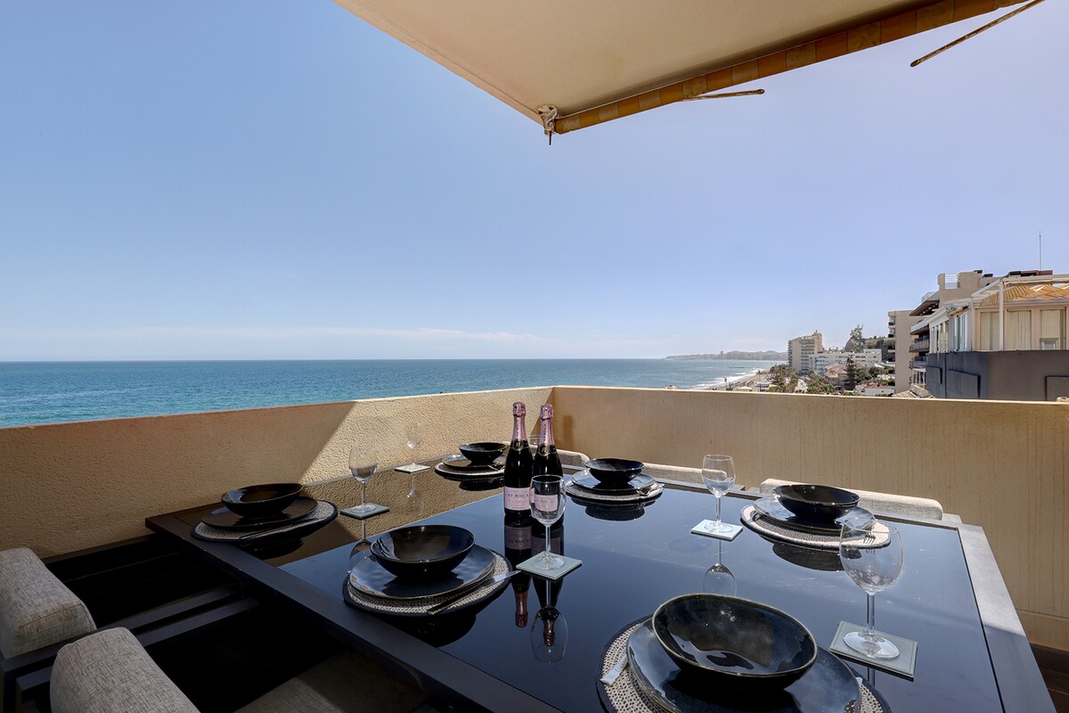 A large outdoor dining table is set with dark plates and sparkling glasses, emphasizing a perfect setting for meals. The table overlooks the beach, with expansive views of the sea and coastline, while the sun casts a bright light on the space.