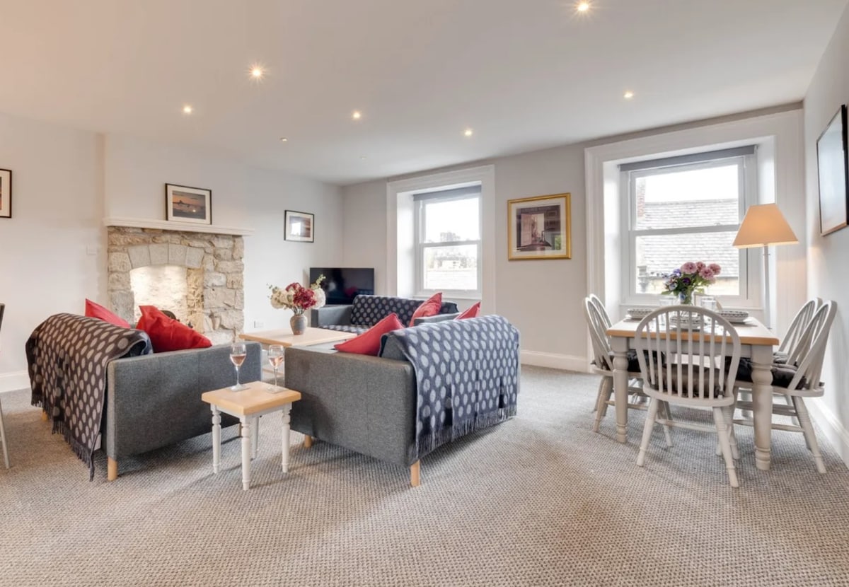 A spacious living area features two cozy sofas adorned with decorative throw blankets. A stone fireplace creates a warm focal point, complemented by large windows letting in natural light. A dining table with light wooden chairs is positioned nearby, providing a comfortable space for meals.