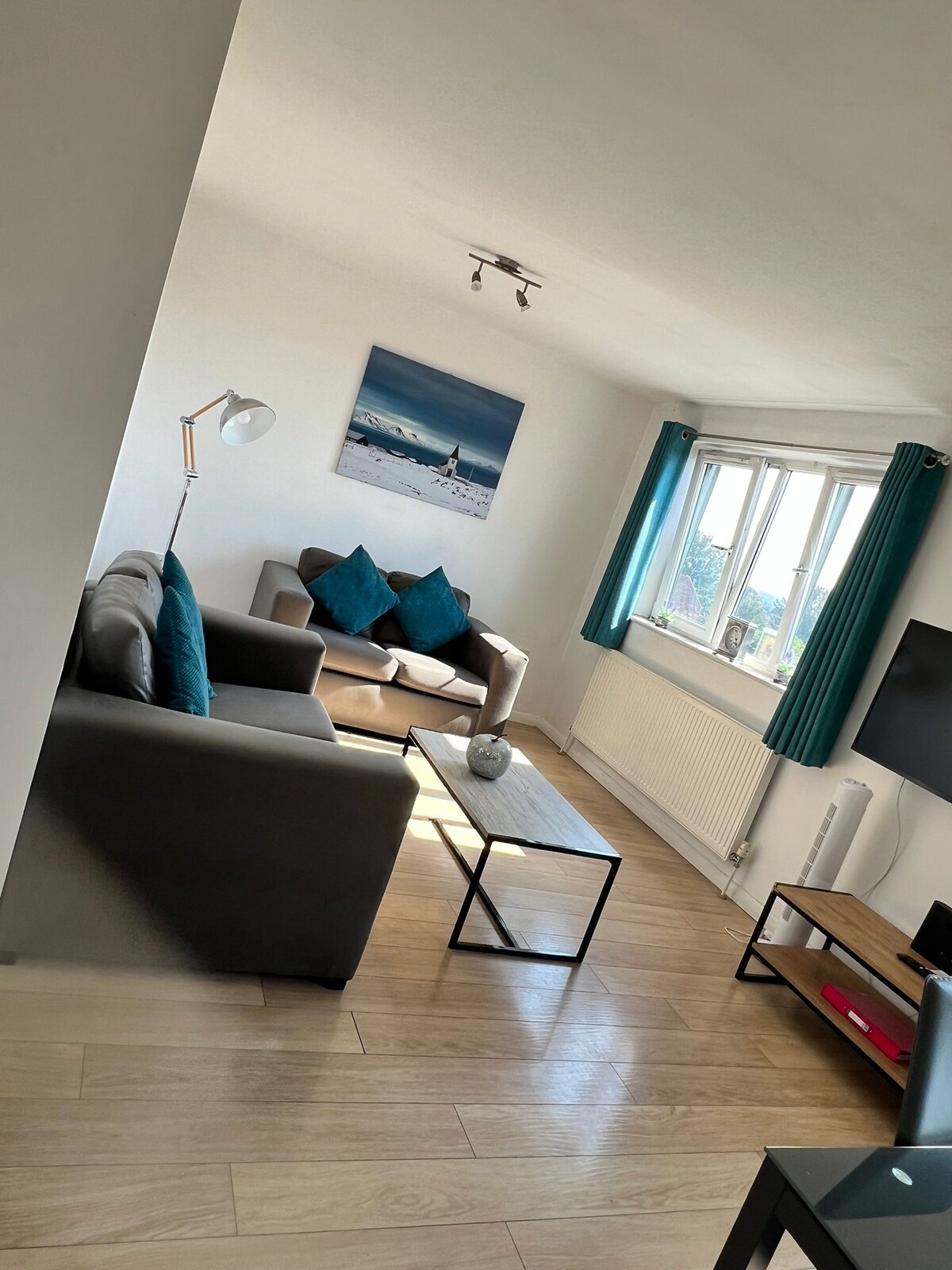 A modern living room features two sofas, one adorned with teal cushions. A minimalist coffee table sits between them, accompanied by a decorative item. Natural light floods the space through large windows, which are dressed with teal curtains. A wall-mounted TV is positioned nearby.