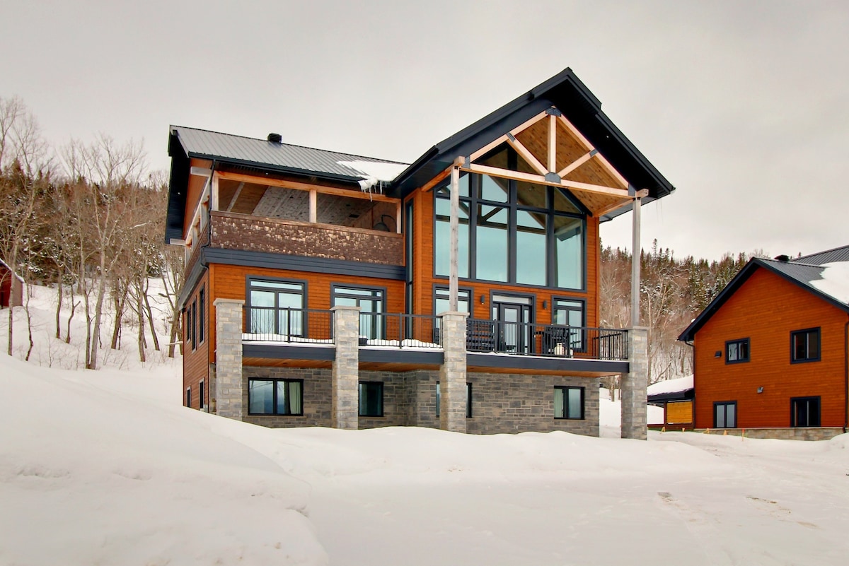 A large chalet is depicted, showcasing a modern architectural design with extensive glass walls. A spacious balcony extends from the upper level, offering views of the surrounding snowy landscape. The exterior features a combination of wood and stone elements, contributing to the rustic yet contemporary aesthetic.